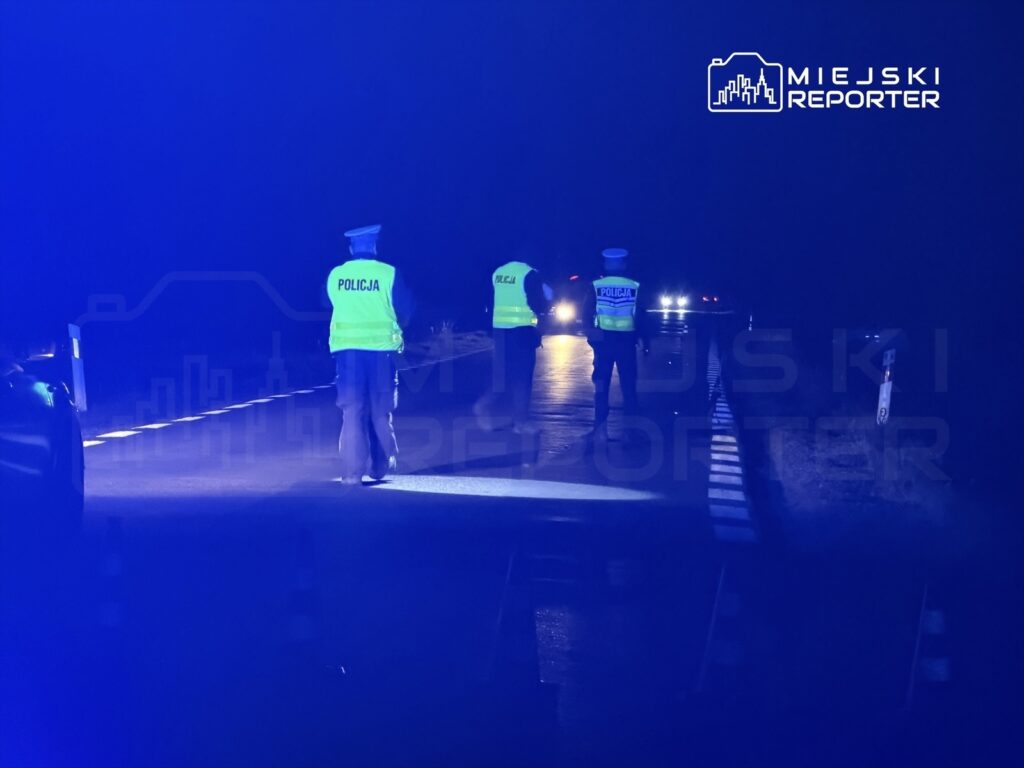 Policjanci w odblaskowych kamizelkach kontrolują ruch na drodze w nocy, z niebieskimi światłami w tle.