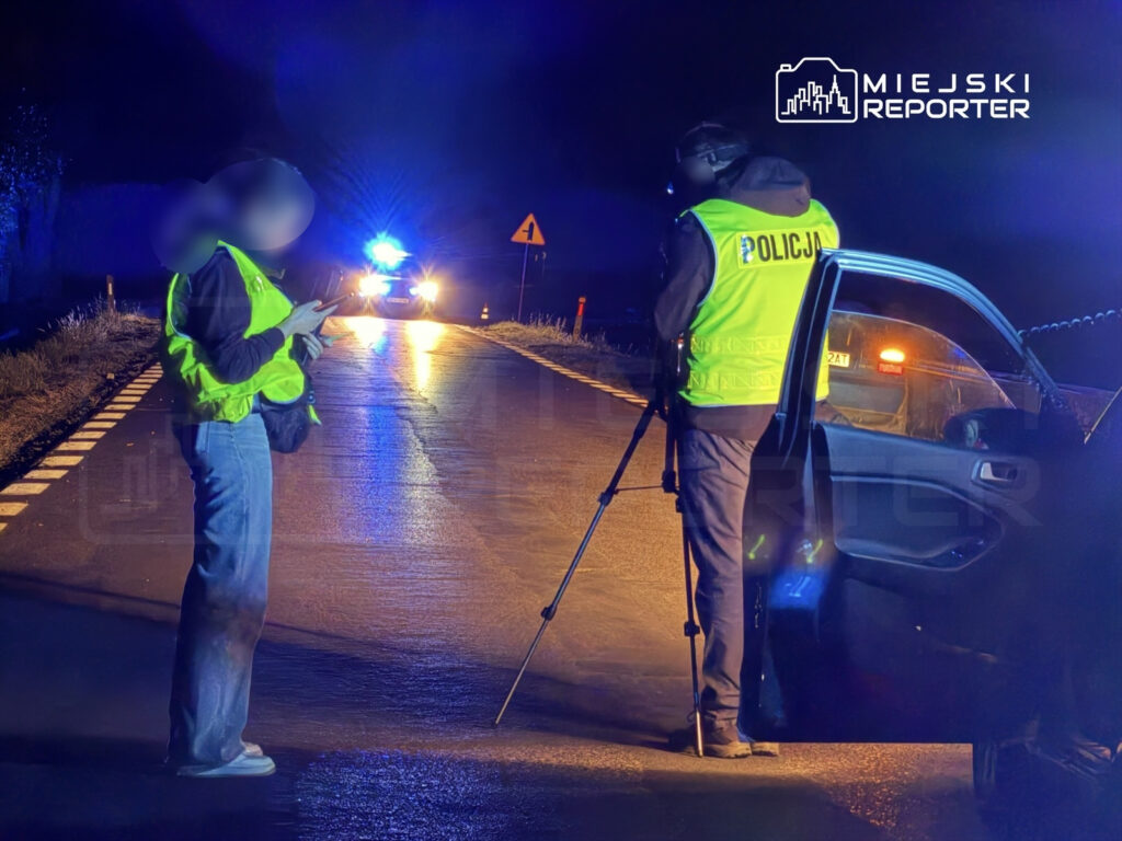 Funkcjonariusz policji rozmawia z osobą w odblaskowej kamizelce na drodze, w tle widać radiowóz z włączonymi światłami.