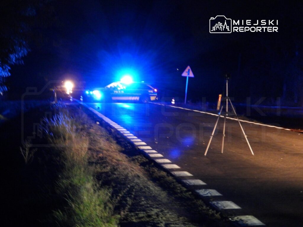 Policyjny radiowóz z niebieskimi sygnalizatorami na drodze, obok stojący statyw z kamerą, nocne oświetlenie.