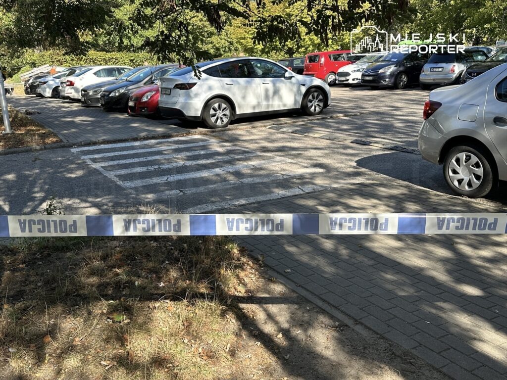 Na parkingu z widocznymi samochodami znajduje się taśma policyjna, oddzielająca część terenu.