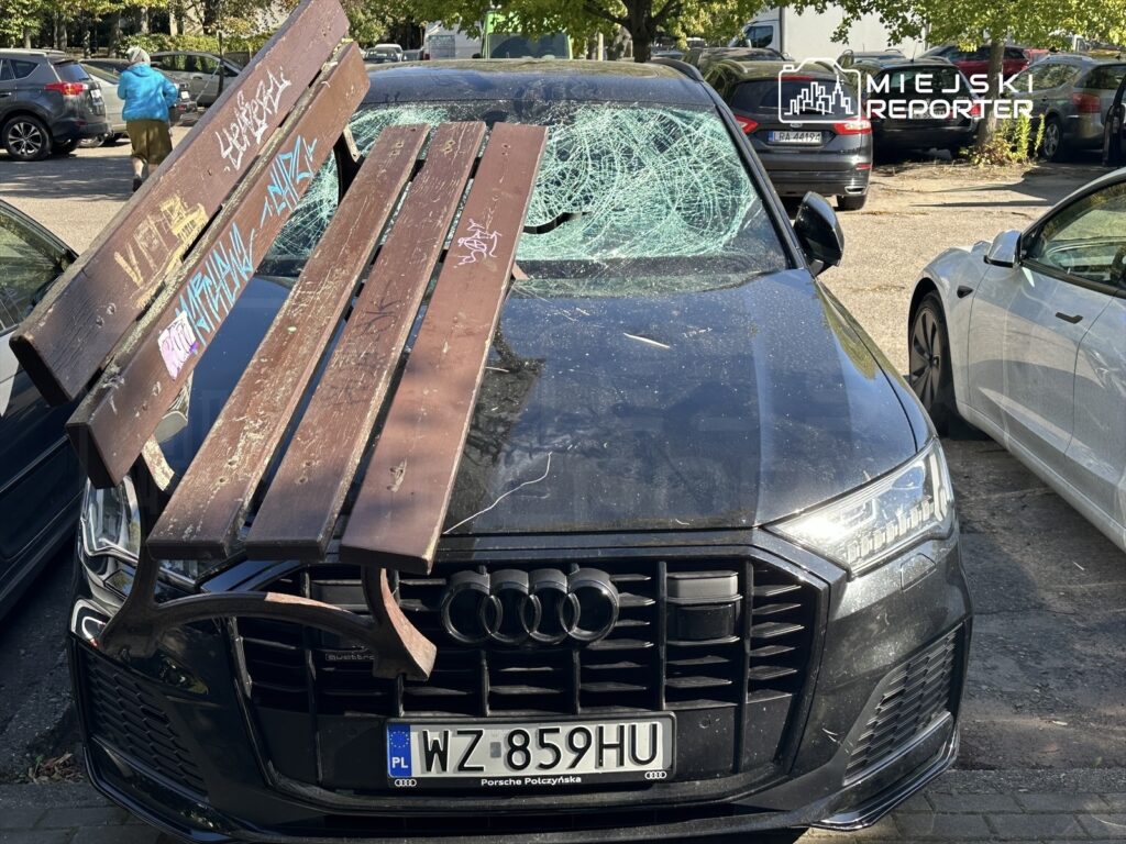 Zniszczony samochód osobowy z rozbitym przednim szkłem, na dachu leży uszkodzona drewniana ławka.