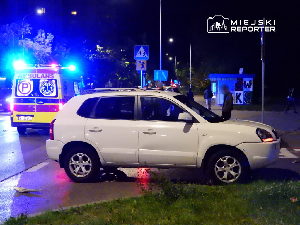 Biały samochód osobowy zatrzymany na skrzyżowaniu, w tle widać ambulans z włączonymi sygnałami świetlnymi.