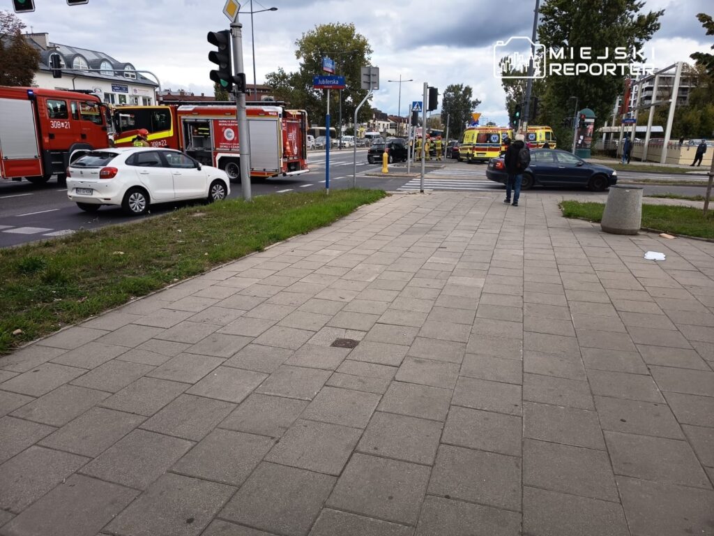Na chodniku stoi mężczyzna z plecakiem, obok zatrzymane pojazdy i w tle widać wozy strażackie oraz karetki.