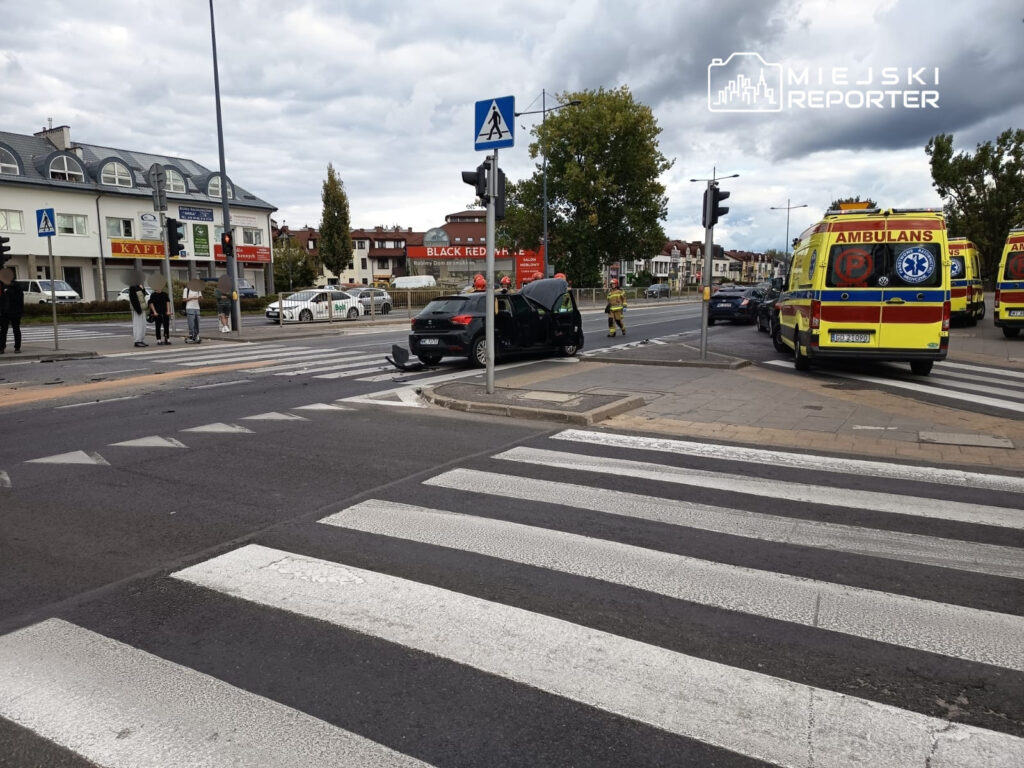Na skrzyżowaniu z sygnalizacją świetlną parkowany jest czarny samochód, obok stoją strażacy i kilka karetek.