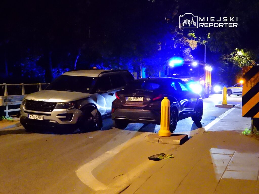 Dwa samochody zaparkowane na drodze, w tle widać pojazd służb ratunkowych z niebieskimi światłami.