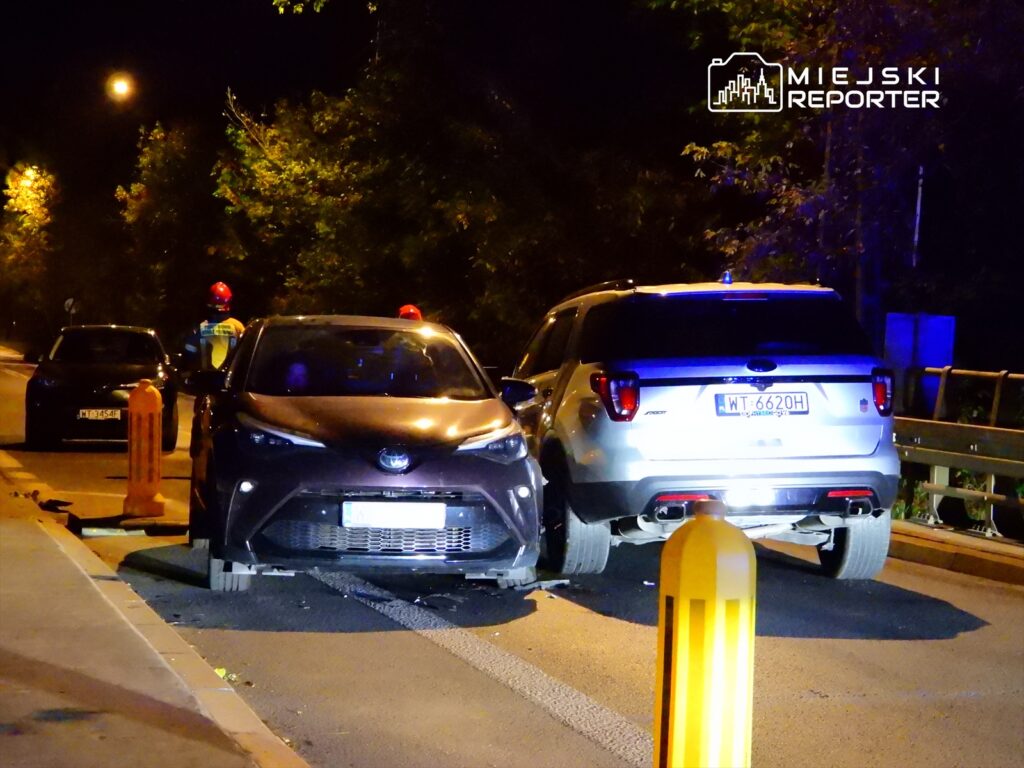 Dwa pojazdy osobowe, jeden czarny i jeden srebrny, zderzone na ulicy w nocy, obok stoją strażacy w kaskach.