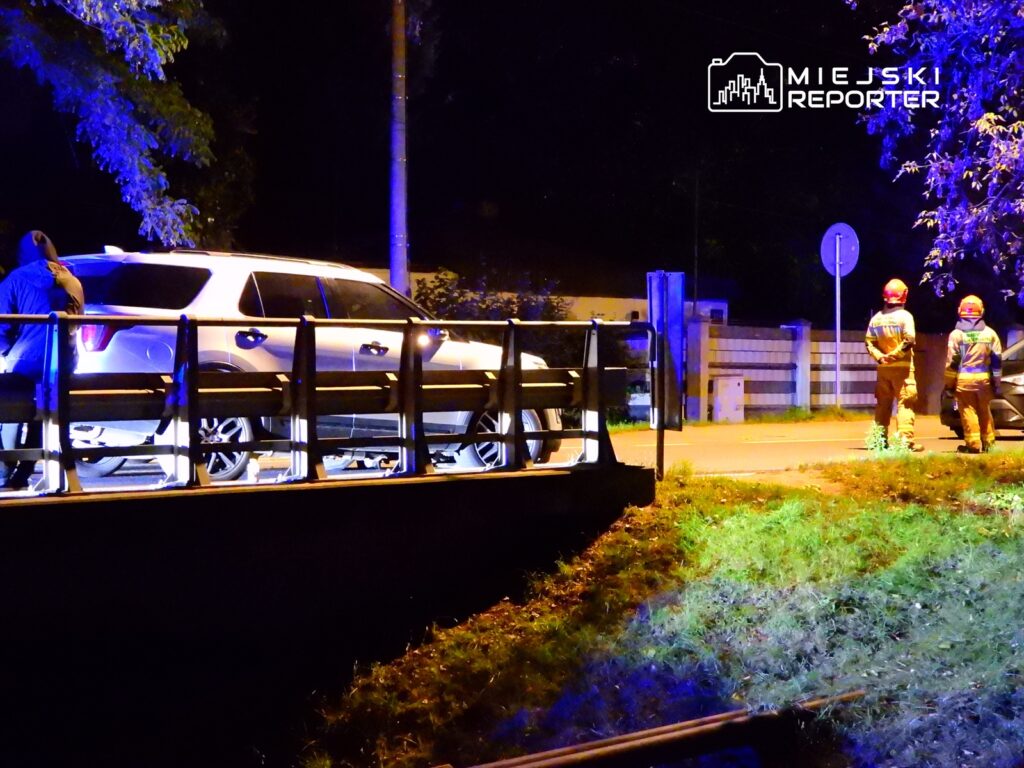 Mężczyzna w ciemnym ubraniu stoi obok białego SUV-a, podczas gdy strażacy w pomarańczowych strojach obserwują sytuację nocą.