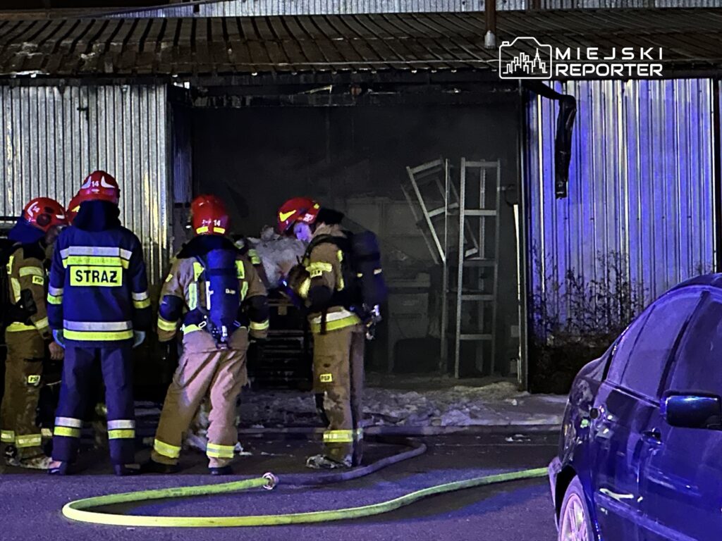 Grupa strażaków w hełmach i mundurach pracuje nad ugaszeniem pożaru w magazynie, obok leży wąż gaśniczy.
