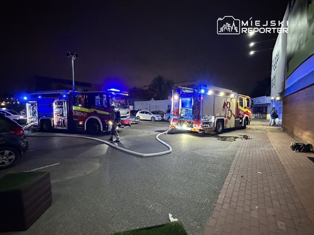 Strażacy w mundurach pracują przy dwóch wozach strażackich na parkingu nocą, z wężami położonymi na ziemi.