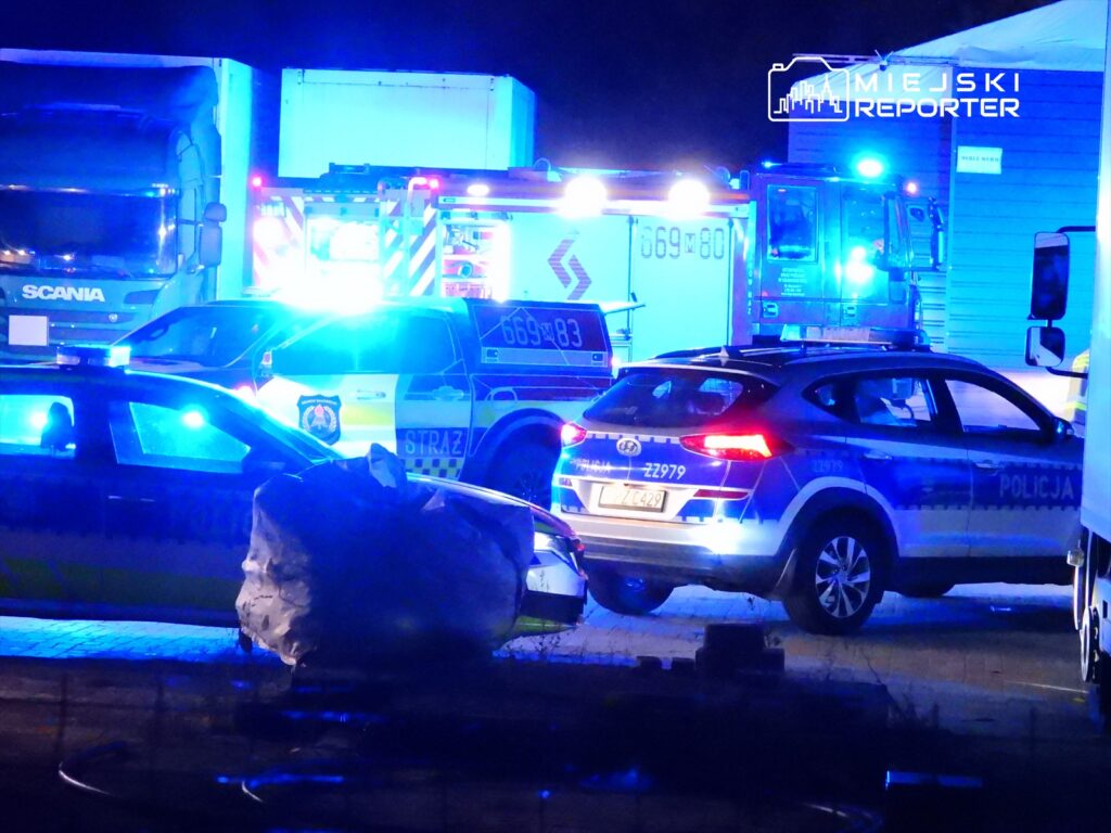 W nocy na miejscu zdarzenia interweniują pojazdy służb ratunkowych, w tym karetka i radiowóz policji, w otoczeniu niebieskich świateł.