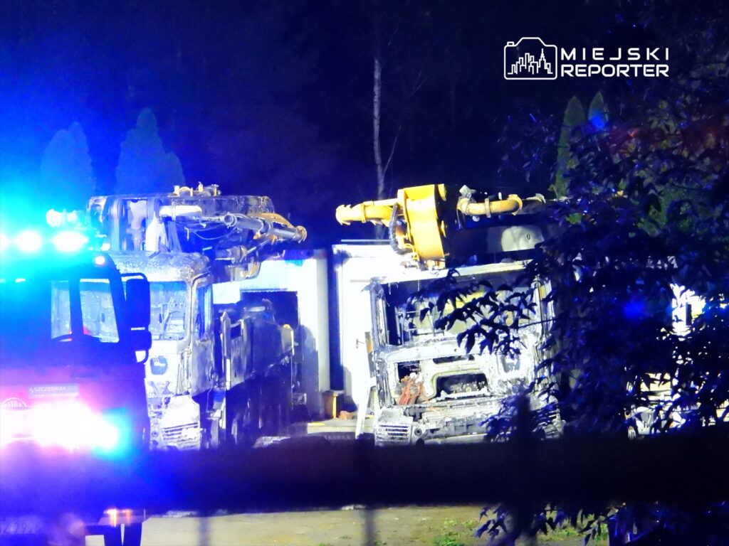 Dwa spalone pojazdy ciężarowe w oświetleniu niebieskich świateł, otoczone przez roślinność nocą.