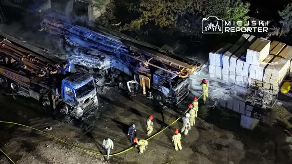 Grupa strażaków w kaskach gasi pożar dwóch zniszczonych ciężarówek na placu budowy, w tle widoczna sterta materiałów budowlanych.