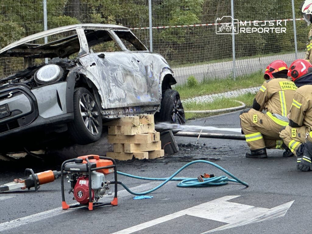 Zniszczony samochód stoi na drewnianych klockach, obok trzech strażaków w mundurach, którzy gaszą pożar.