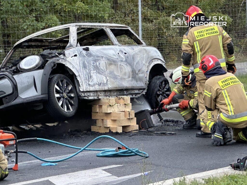 Czterech strażaków pracuje nad usunięciem wraku spalonego samochodu na drodze, używając sprzętu ratunkowego.