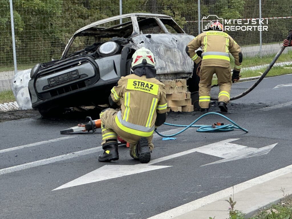 Strażacy gaszą płonący samochód osobowy na poboczu drogi, używając węża strażackiego i narzędzi.