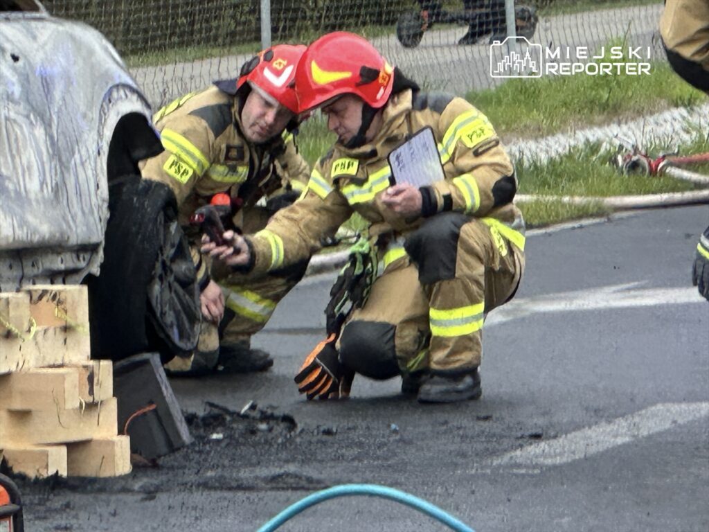 Dwóch strażaków w kaskach bada uszkodzone koło samochodu, otoczeni przez sprzęt gaśniczy na drodze.