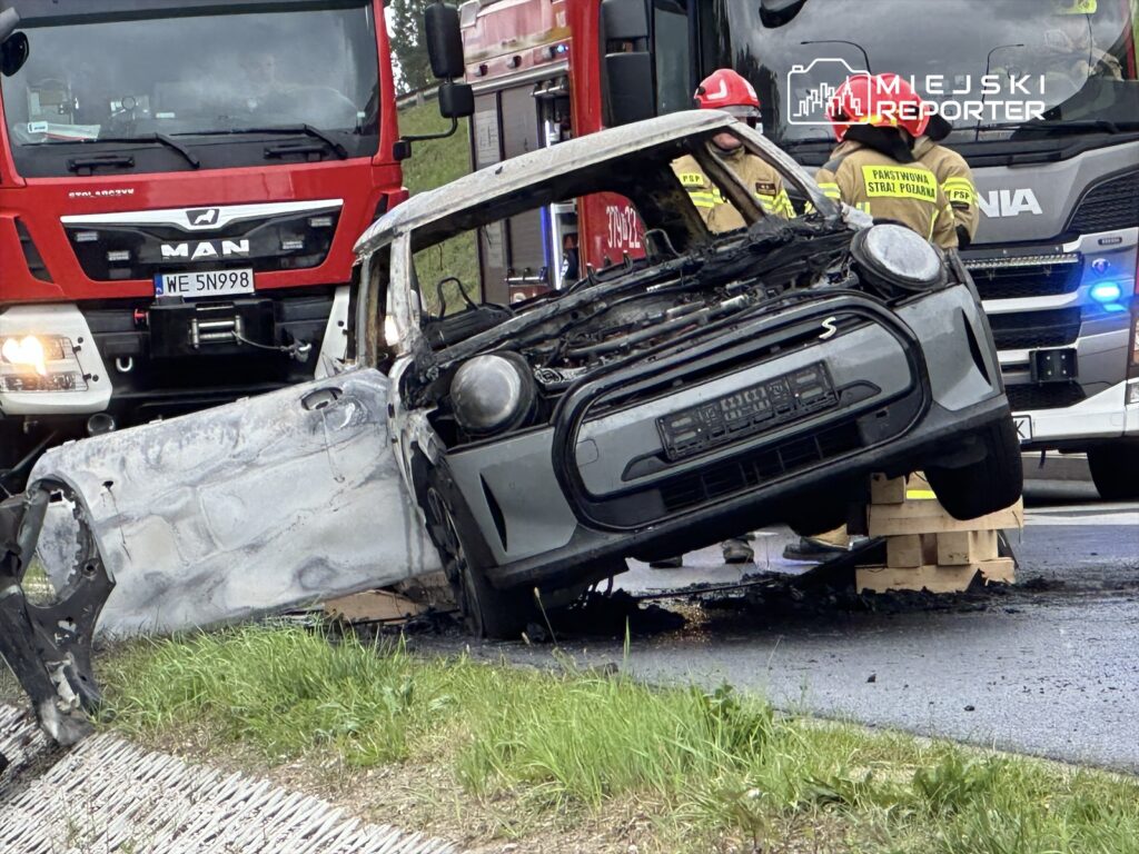 Spalony samochód stoi na poboczu drogi, obok działają strażacy i woz strażacki.