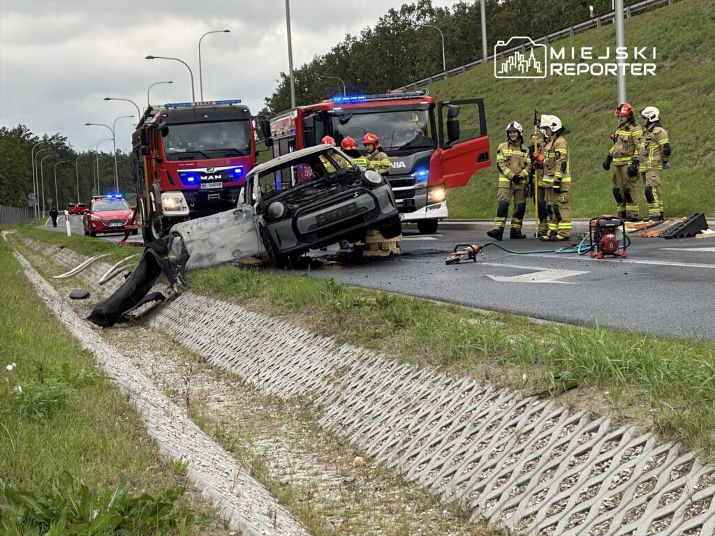 Wielu strażaków pracuje przy przewróconym samochodzie na poboczu drogi, obok widać wozu strażackie z włączonymi sygnałami.