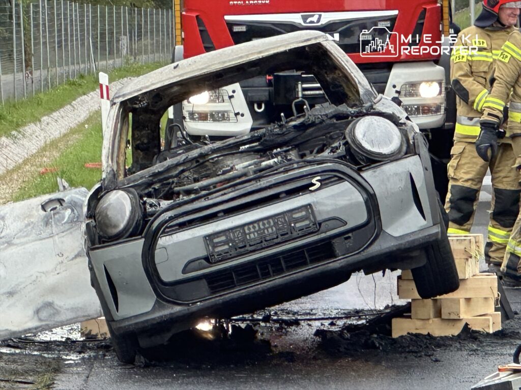 Spalony samochód osobowy leży na drewnianych paletach obok drogi, a w tle widoczni strażacy w mundurach.