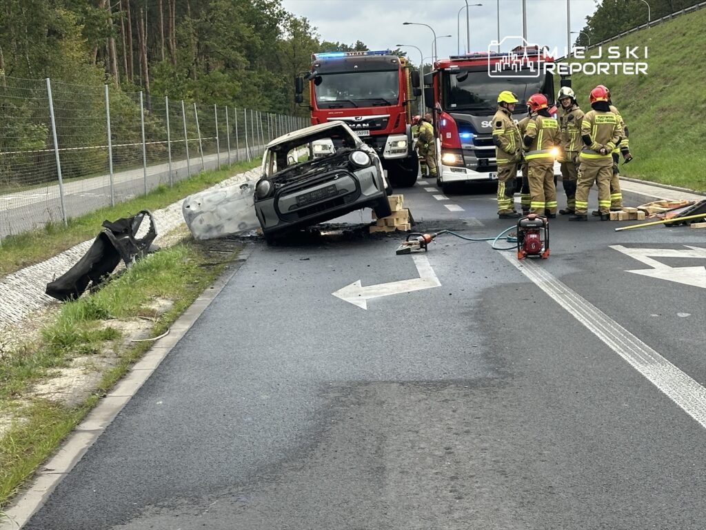 Wielu strażaków pracuje przy przewróconym samochodzie na drodze, obok stoją wozu strażackie.