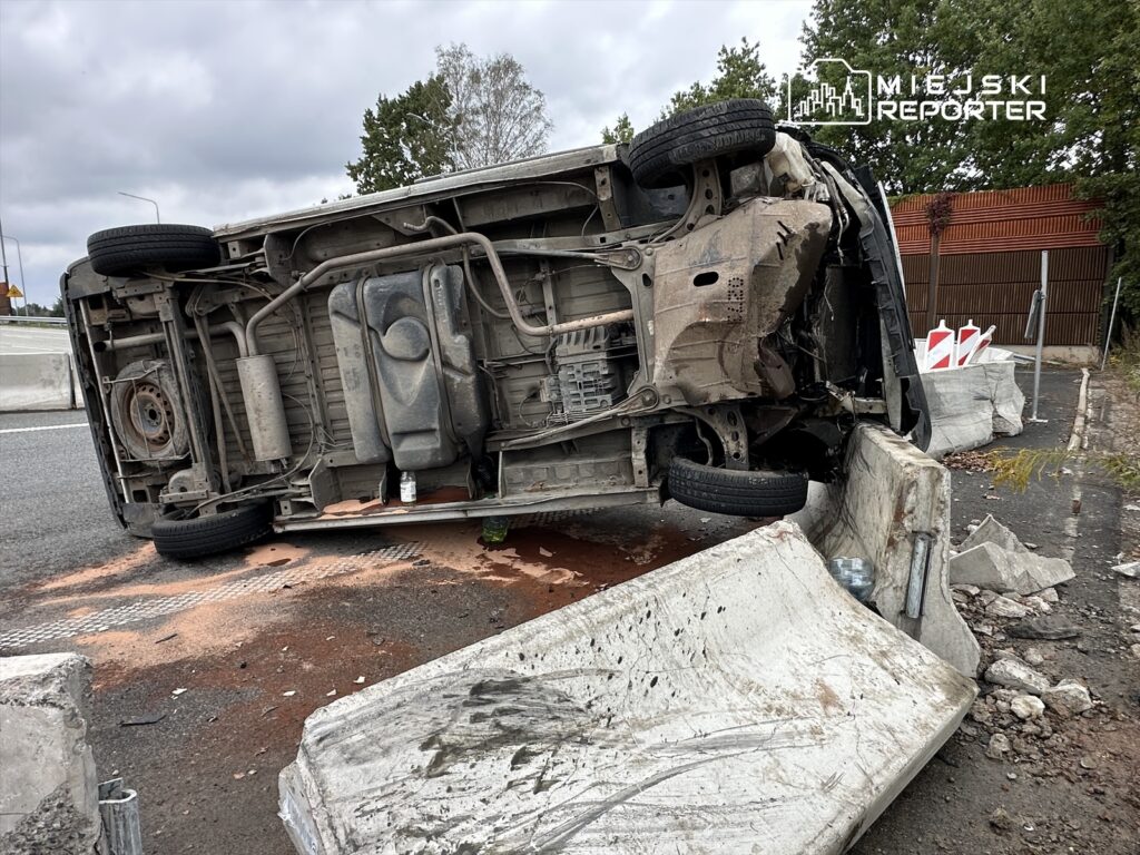 Odwrócone auto osobowe leży na boku obok betonowych bloków, z plamą płynu na jezdni.