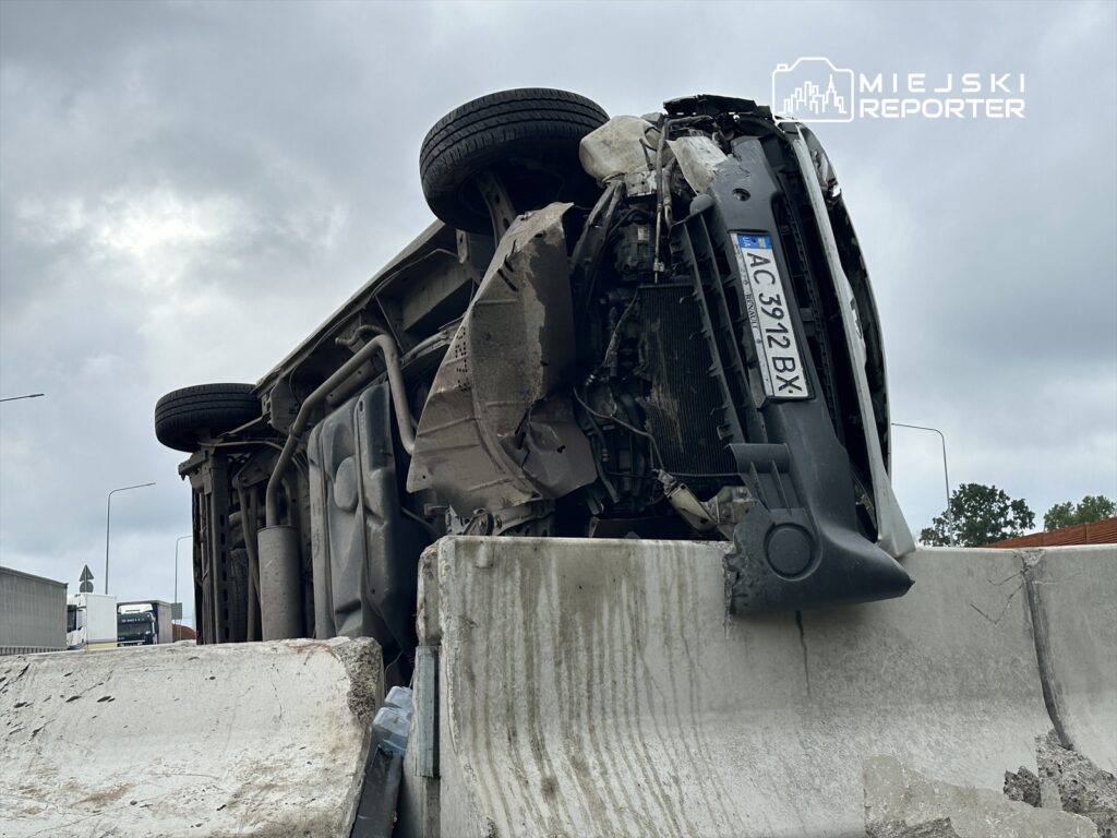 Przewrócone auto na barierce w miejskim otoczeniu, z widocznymi uszkodzeniami i zarysowaniami.