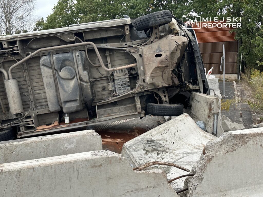 Przewrócony samochód osobowy leży na betonowych blokach w zarośniętym otoczeniu.