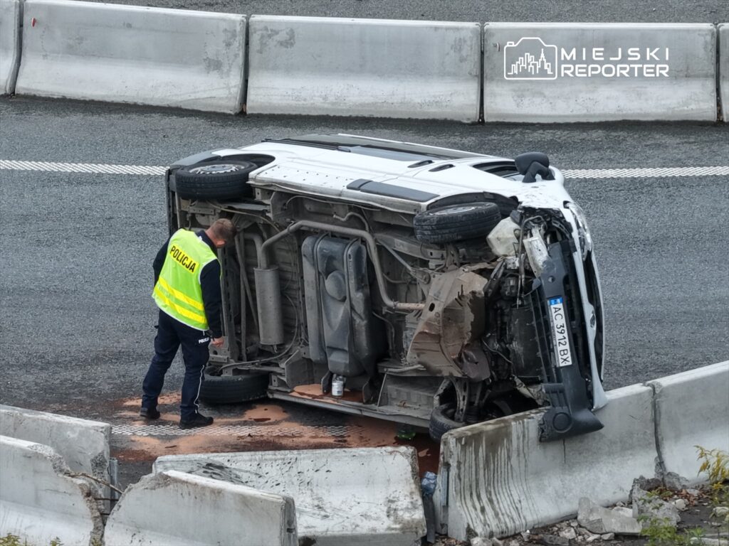 Policjant w odblaskowej kamizelce bada przewrócony samochód na drodze, obok betonowe bariery.