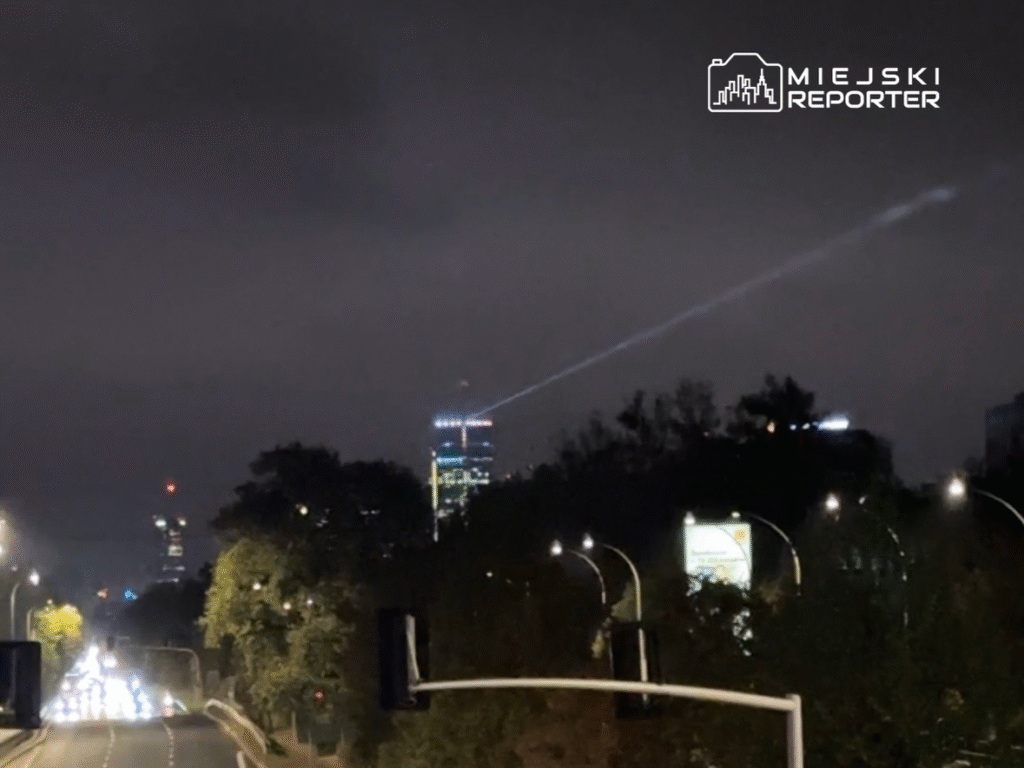 Na tle nocnego nieba widać wieżowce, z jednego z nich emanuje smuga światła. Ulica wypełniona jest ruchem samochodowym.