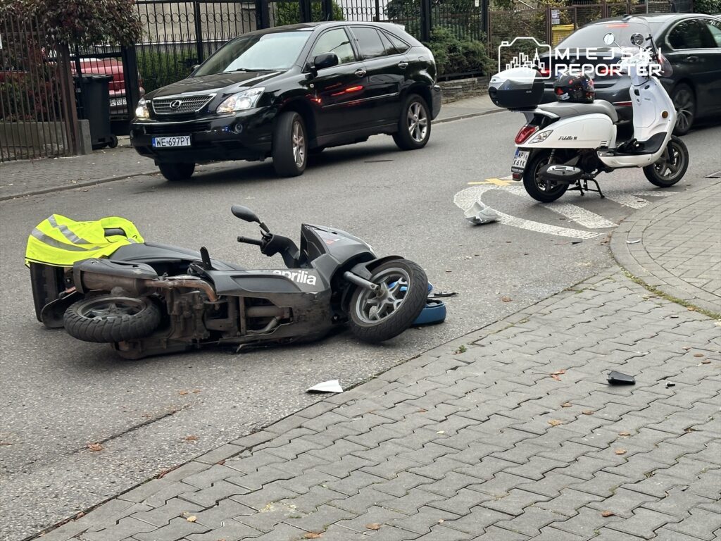 Przewrócony skuter leży na jezdni obok białego skutera, w tle zaparkowane samochody.