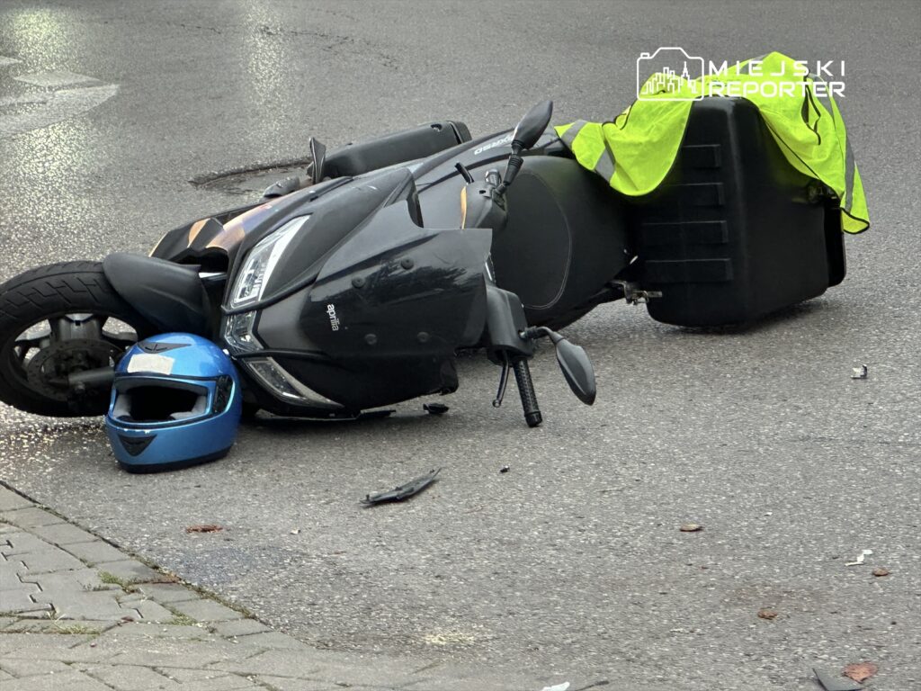 Skuter leży na boku na jezdni, obok niego niebieski kask oraz żółta kamizelka odblaskowa.