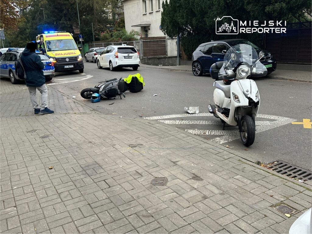 Mężczyzna w ciemnej kurtce obserwuje wypadek motocyklowy na ulicy, obok leży przewrócony skuter.