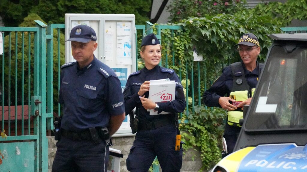 Trzech policjantów w mundurach spaceruje obok zielonego ogrodzenia, jedna z kobiet trzyma dokumenty i uśmiecha się.