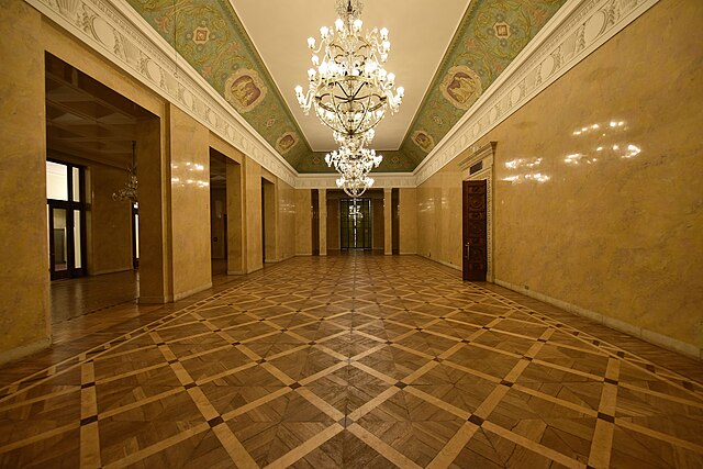 Przestronna sala z parkietem w jodełkę, ozdobiona żyrandolami i malowanymi sufitami.