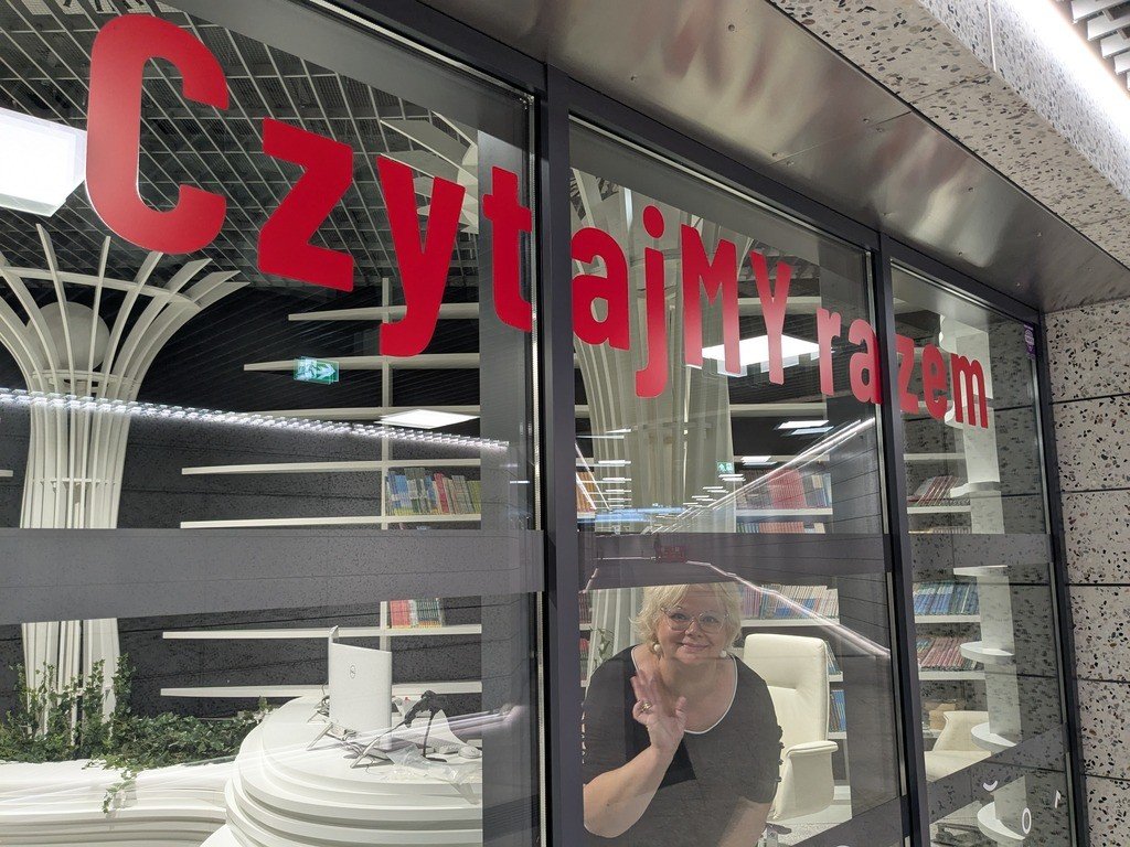 Kobieta z okularami macha ręką przez szklane drzwi nowoczesnej biblioteki z napisem "CzytajMy razem".