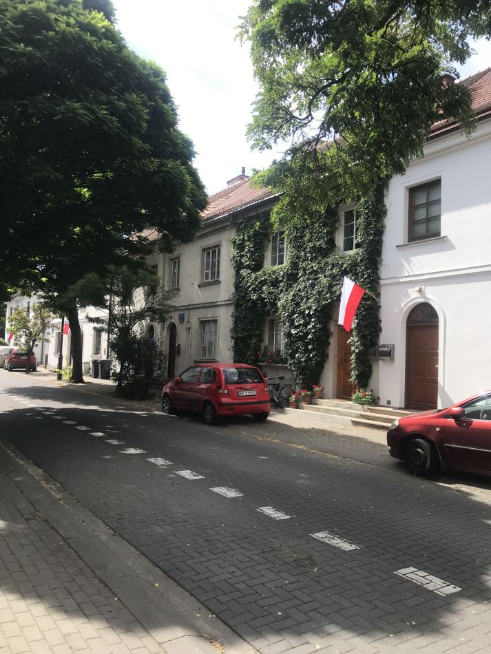 Czerwony samochód zaparkowany na ulicy obok budynków z zielonymi pnączami i flagami Polski na elewacji.