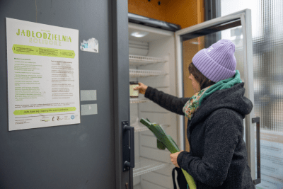 Kobieta w fioletowej czapce wkłada jedzenie do lodówki w jadłodzielni, obok znajduje się informacyjny plakat.