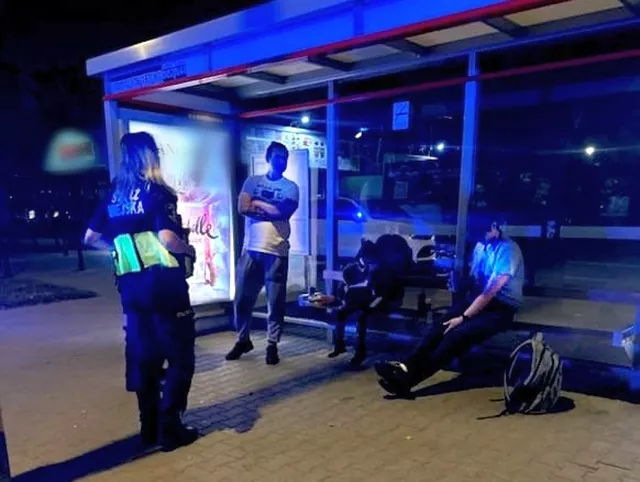 Funkcjonariusz policji rozmawia z grupą młodych ludzi na przystanku autobusowym w nocy.