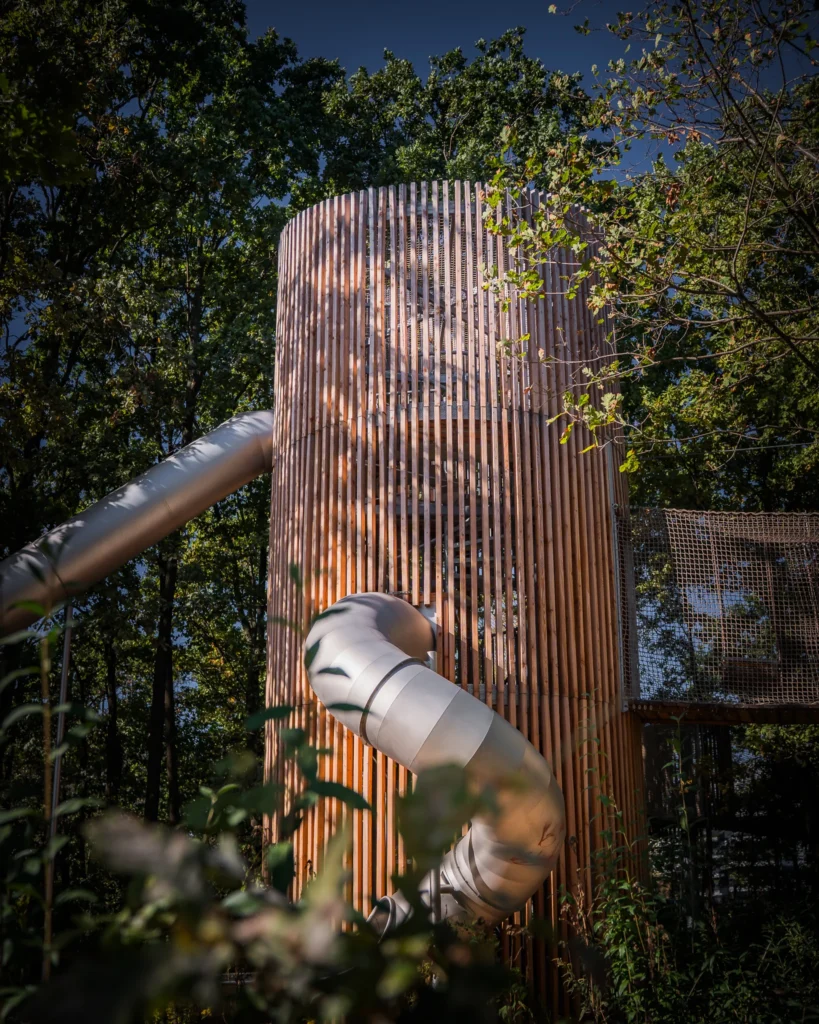 Nowoczesny plac zabaw z drewnianą wieżą i metalową zjeżdżalnią, otoczony zielenią drzew i krzewów.