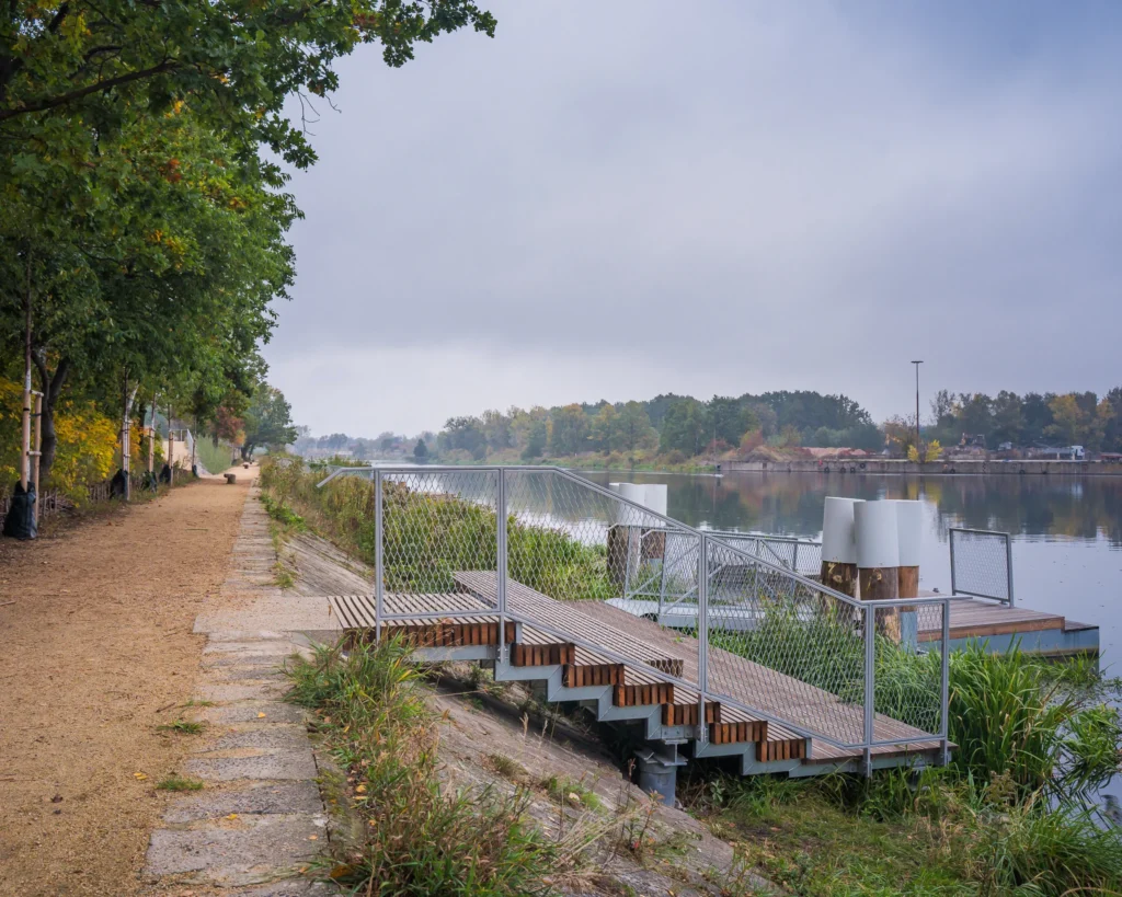 Ścieżka spacerowa wzdłuż rzeki, z drewnianym pomostem i zielenią w tle, pod pochmurnym niebem.