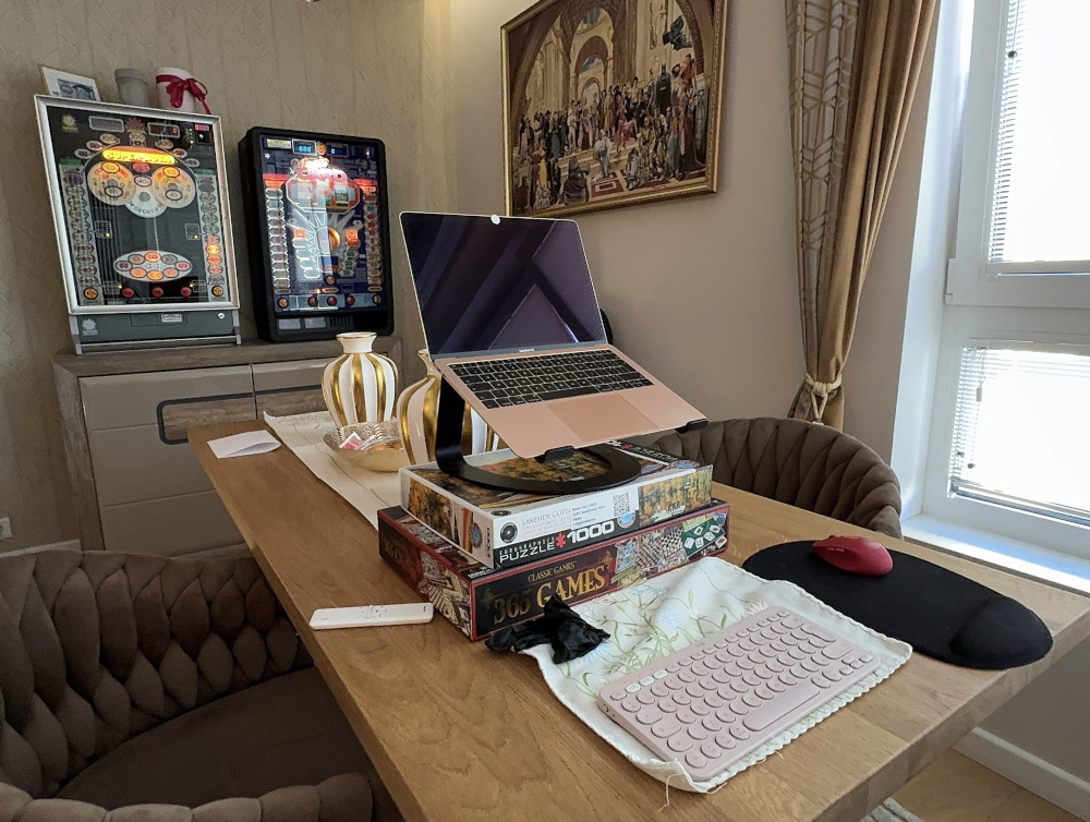 Laptop umieszczony na stosie gier planszowych, obok myszka komputerowa i różowa klawiatura, w jasnym wnętrzu z automatami do gier.