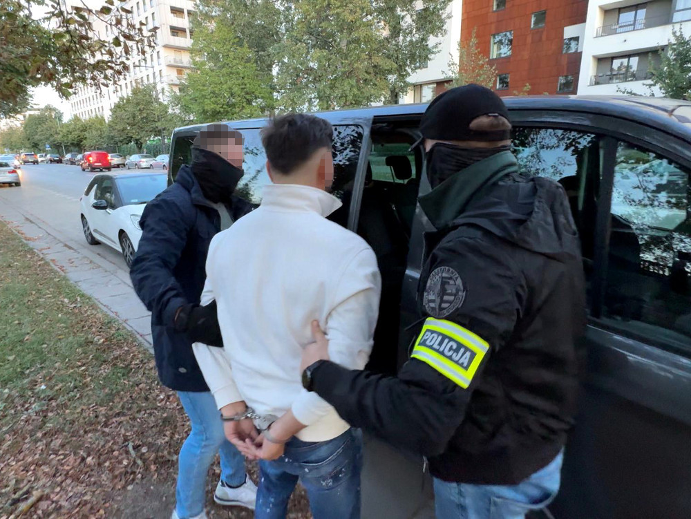 Mężczyzna w białej bluzie jest zatrzymywany przez dwóch policjantów w czarnych maskach na ulicy w mieście.