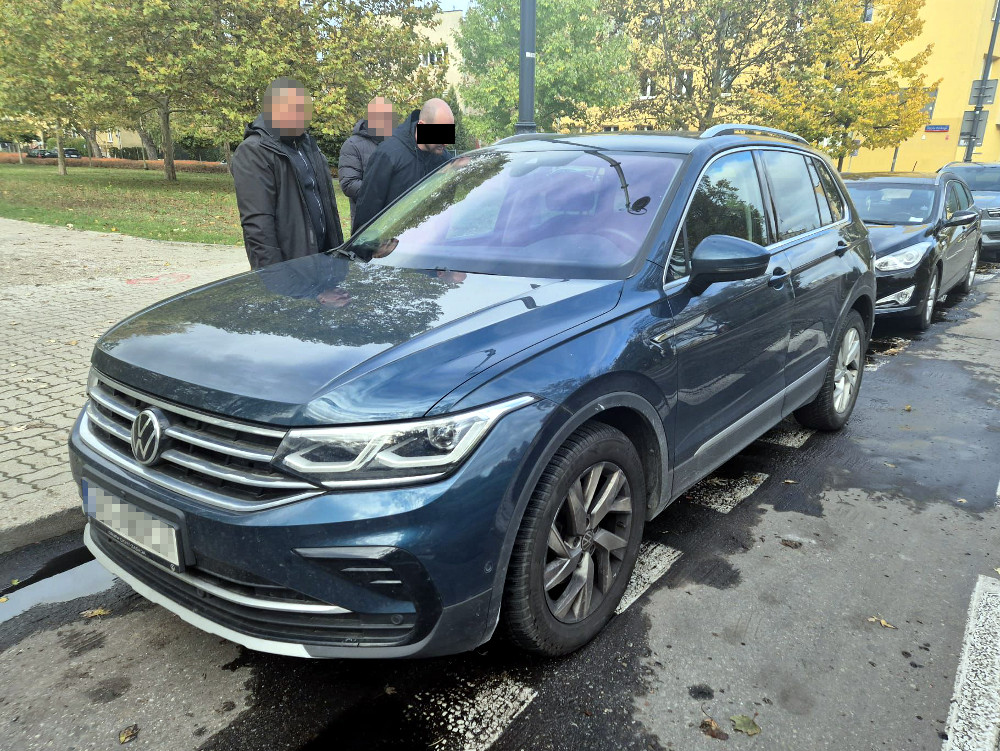 Trzech mężczyzn rozmawia obok niebieskiego samochodu marki Volkswagen zaparkowanego na ulicy, w tle drzewa.