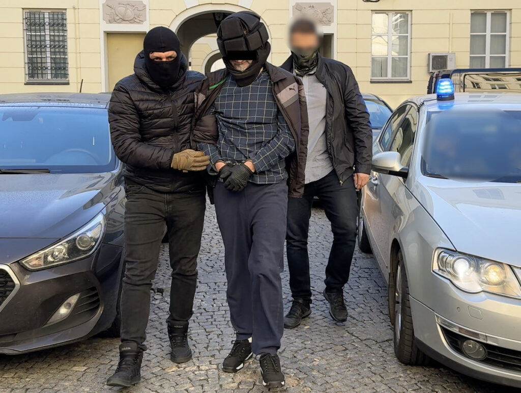 Mężczyzna w kajdankach prowadzony przez dwóch uzbrojonych funkcjonariuszy w pobliżu zaparkowanych samochodów.