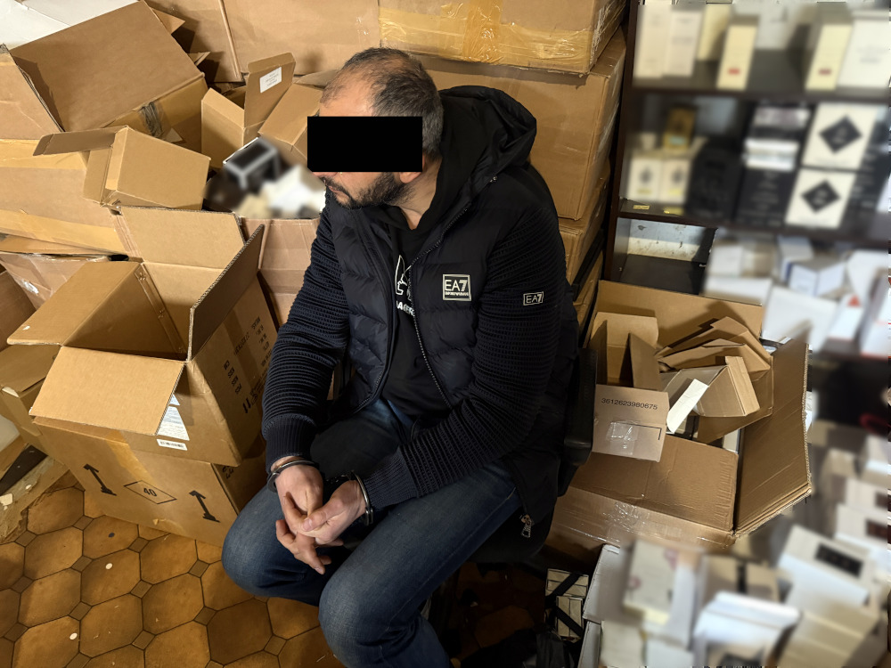 Mężczyzna w czarnej kurtce siedzi na krześle wśród rozpakowanych kartonów w magazynie.