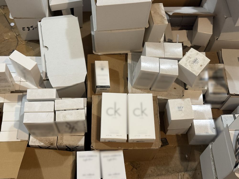 Zestaw opakowań kosmetyków i perfum ułożonych na podłodze w magazynie, wśród kartonów i pudełek.