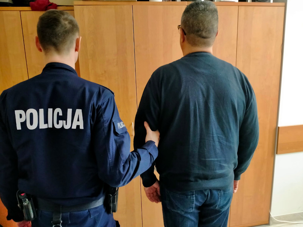 Policjant w mundurze prowadzi mężczyznę w ciemnym swetrze w kierunku szafek w pomieszczeniu.