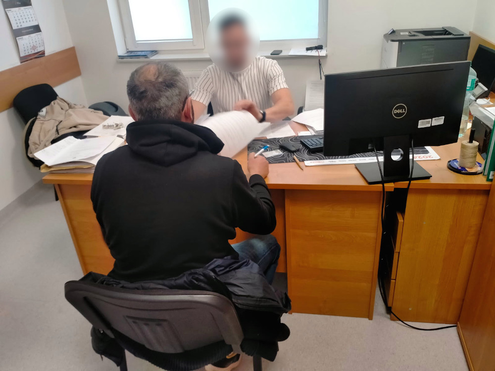 Mężczyzna w czarnej bluzie siedzi na krześle, rozmawiając z mężczyzną w koszuli przy biurku w biurze.