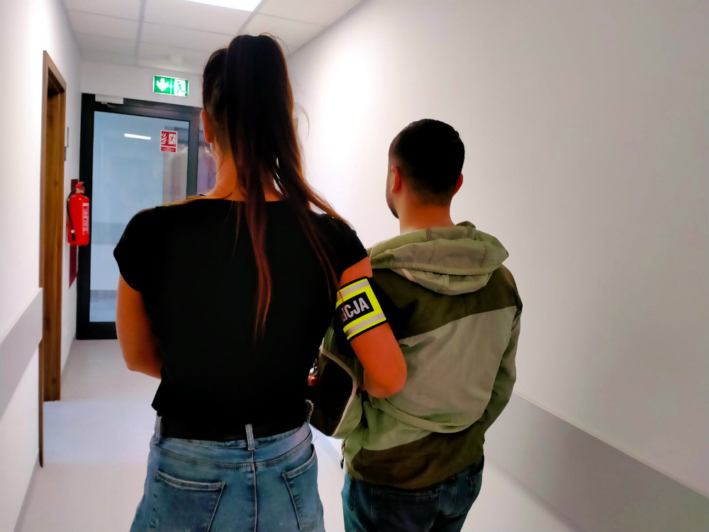 Kobieta w czarnej koszulce z opaską "POLICJA" prowadzi mężczyznę w szarej bluzie przez korytarz szpitalny.