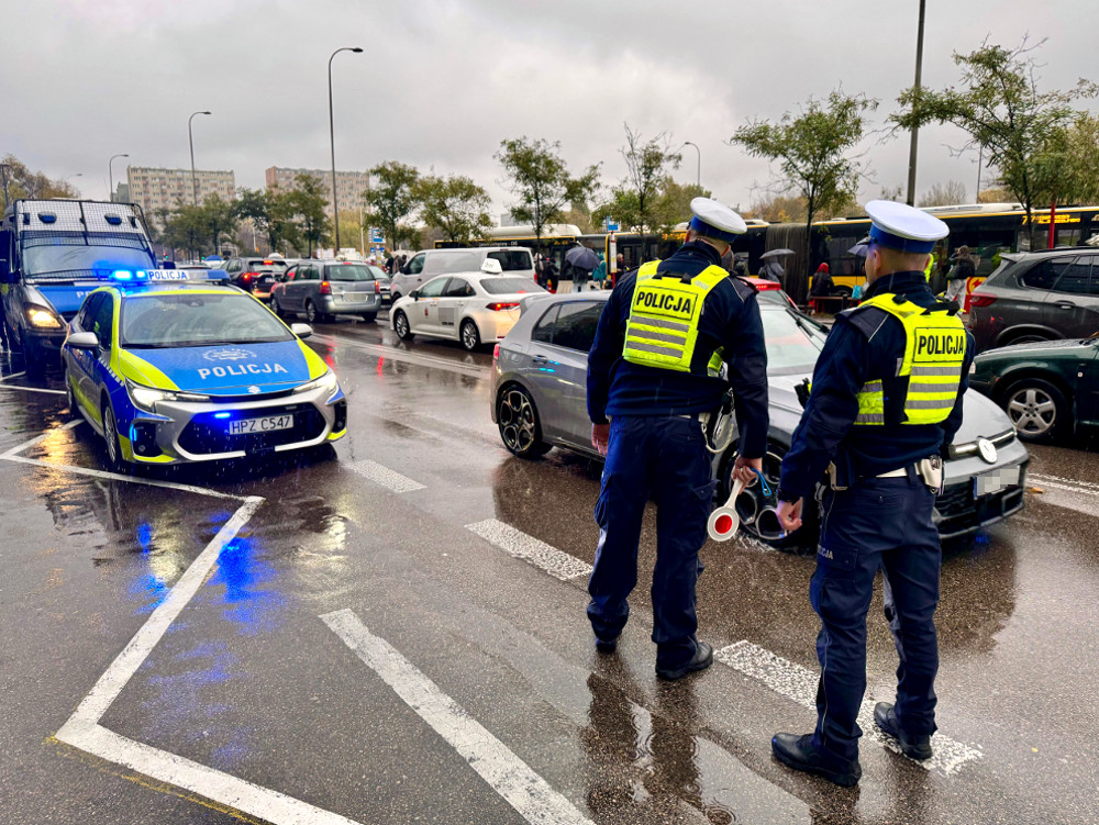 Dwóch policjantów w mundurach kontroluje ruch na zatłoczonej ulicy podczas deszczu, obok radiowóz z włączonymi sygnałami.
